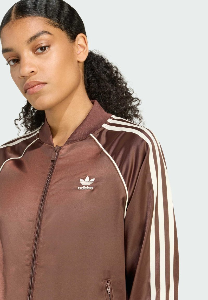 Brown satin zip-up jacket featuring white stripes on shoulders and a small white logo on the left chest, with a ribbed collar and smooth texture.