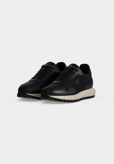 Paire de baskets noires à lacets avec semelles blanches et semelles extérieures noires texturées, présentée sur un fond clair uni.