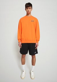 Felpa arancione brillante con polsini a coste, abbinata a pantaloni corti neri. Il modello indossa calze bianche e sneakers crema con suola chiara.
