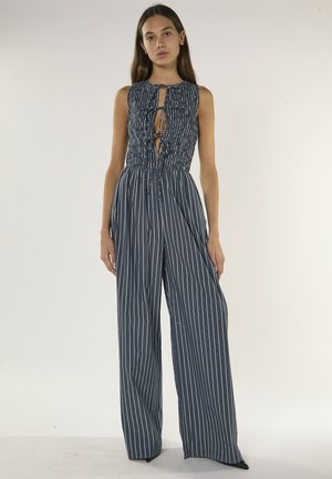 Frau trägt einen ärmellosen marineblauen Jumpsuit mit vertikalen weißen Streifen und vorderen Bindedetails, stehend vor einem schlichten hellen Hintergrund.