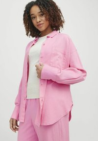 Roza lanena srajca z dolgimi rokavi, žepom na levi strani in zapenjanjem na gumbe, v kombinaciji z svetlejšim zgornjim delom in usklajenimi hlačami.