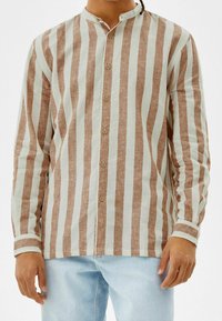 Långärmad skjorta med vertikala ränder i ljusbrun och off-white, tillverkad av ett ventilerande tyg. Knäppning framtill med ståkrage.