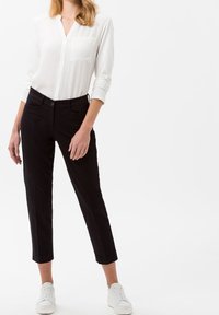 Camicia bianca a maniche lunghe con bottoni e taschino sul petto, abbinata a pantaloni neri su misura che si stringono alla caviglia, indossata con sneaker bianche.