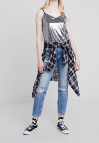 Femme portant un débardeur en sequins argentés, un jean bleu déchiré, une chemise à carreaux nouée autour de la taille et des baskets montantes noires sur fond uni.