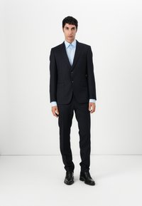 Marineblauer Anzug mit taillierter Passform, ausgestattet mit zwei Knöpfen, Reverskragen und seitlichen Taschen. Getragen über einem hellblauen Hemd, kombiniert mit schwarzen Schuhen.