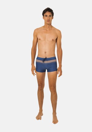 Jeune homme debout pieds nus portant un short de bain bleu avec des rayures horizontales marron clair et un cordon à l'avant.