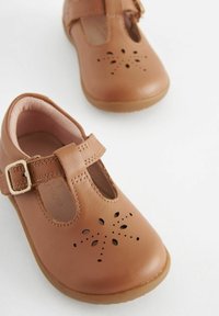 Brązowe skórzane buty z designem T-strap, perforowanymi kwiatowymi wzorami oraz zapięciem na klamrę. Miękka podszewka wewnętrzna i zaokrąglony czubek.
