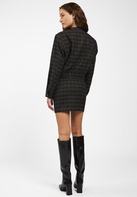 Vrouw die wegkijkt in een donker geruite blazer en bijpassende mini-rok met zwarte kniehoge haklaarzen op een witte achtergrond.