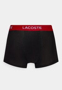 Fekete boxer alsónadrág piros elasztikus derékpánttal, melyen a "LACOSTE" logó látható. Az anyag sima, az alsónadrág testhezálló kialakítású, és nincsenek látható minták.