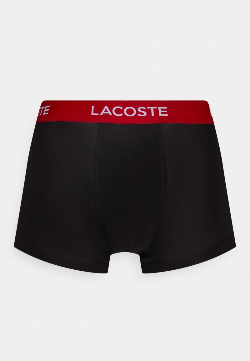 Zwarte boxershorts met een rode elastische tailleband met het "LACOSTE" logo. De stof is glad met een aangesloten ontwerp en zonder zichtbare patronen.