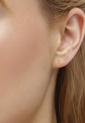 Nahaufnahme eines Ohrs mit einem goldenen Ohrstecker in Form des Buchstabens "G" bei einer Person mit heller Haut und blondem Haar.