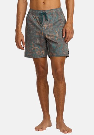 Männliches Modell trägt türkis-braune Badehose mit Blumenmuster und Kordelzug an der Taille, steht barfuß vor einem schlichten Hintergrund.