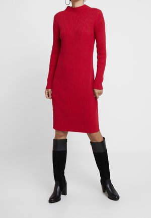 Femme portant une robe midi en maille côtelée rouge à manches longues, associée à des bottes noires montantes à talons, se tenant devant un fond uni.