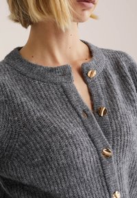 Grå stickad kofta med rund halsringning, strukturerat tyg och guldfärgade knappar som stängning. Detaljerad med ett ribbstickat mönster.