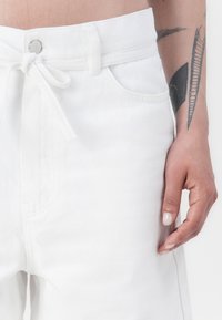 Hvit denim shorts med snor i midjen, frontlommer og en glatt tekstur. Har en sølvknapp og synlige sømdetaljer.