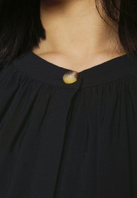 Svart blus med veck, som har en stor, glänsande gul knapp vid halsringningen, vilket framhäver dess textur och design detaljer.