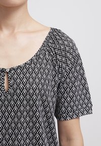 Blus med svart och vitt mönster som har rund halsringning med en nyckelhålsdetalj och korta ärmar, tillverkad av lättviktig tyg.