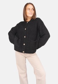 Schwarze, strukturierte Strickjacke mit einer lässigen Passform, ausgestattet mit goldenen Knöpfen, großen Vorderaschen und abgesenkten Schultern, kombiniert mit hellen Hosen.