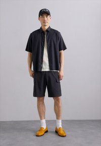 Čierna košeľa s krátkym rukávom na gombíky cez biely tričko, v kombinácii s tmavými cargo šortkami a oranžovými mokasínami, doplnené bielymi ponožkami.