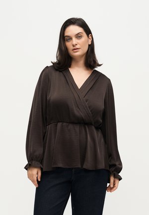 Femme aux cheveux bruns mi-longs portant un chemisier cache-cœur à manches longues brun foncé et un pantalon foncé, debout devant un fond clair uni.