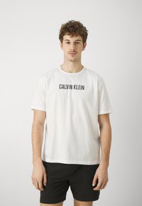 Biała bawełniana koszulka z logo "CALVIN KLEIN" w czarnym kolorze na przodzie, okrągły dekolt, luźny krój, noszona z czarnymi szortami.