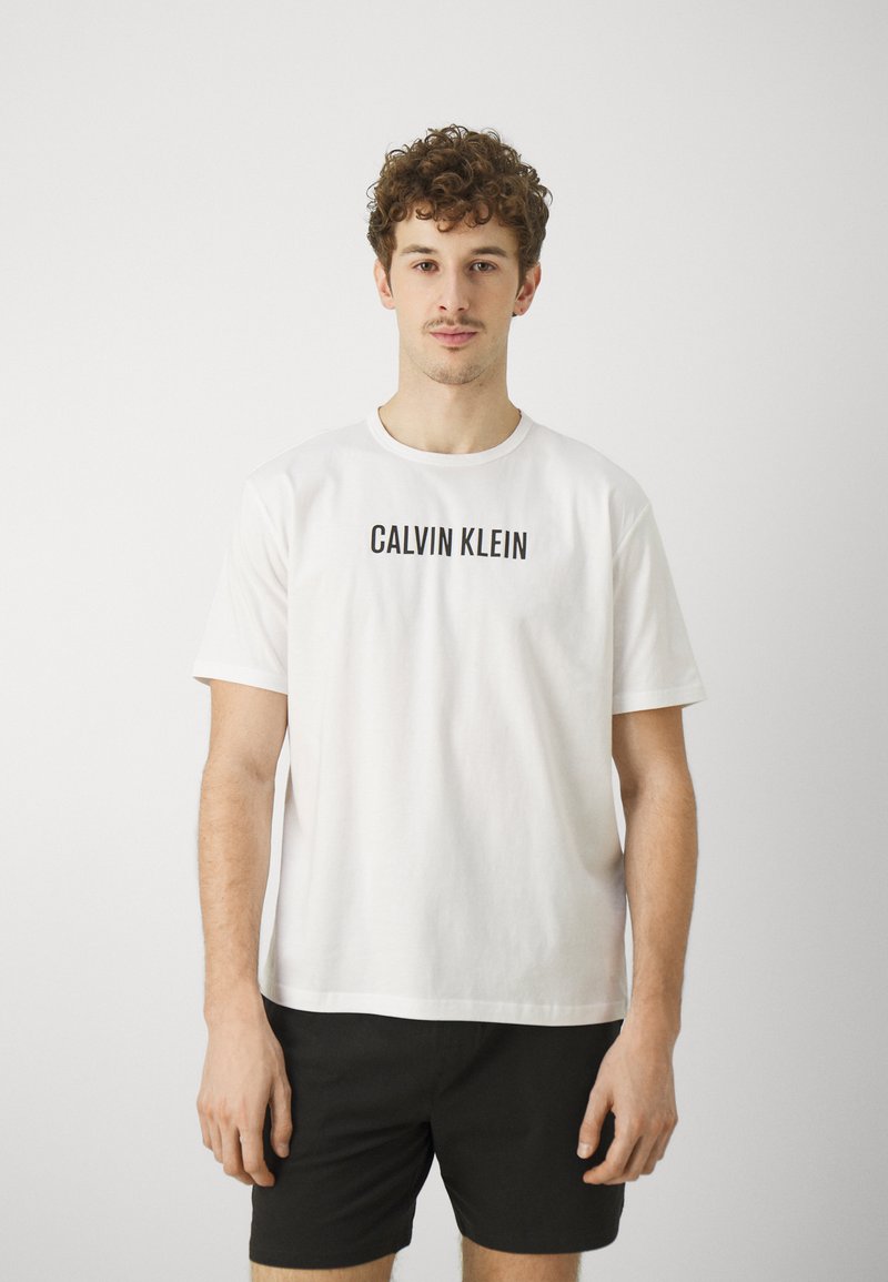 Biała bawełniana koszulka z logo "CALVIN KLEIN" w czarnym kolorze na przodzie, okrągły dekolt, luźny krój, noszona z czarnymi szortami.