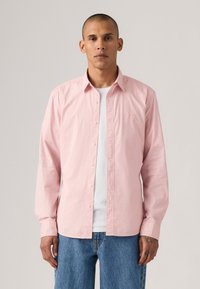 Camisa de botones de manga larga rosa, fabricada en algodón, corte entallado, con un bolsillo en el pecho, usada sobre una camiseta blanca, combinada con jeans azules.