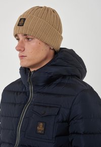 Giacca imbottita navy con colletto alto e chiusura zip, dotata di taschino sul petto e berretto lavorato a coste beige.