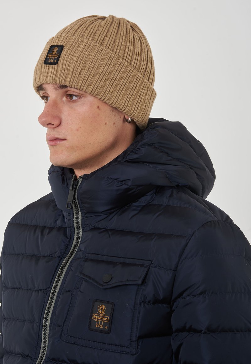Giacca imbottita navy con colletto alto e chiusura zip, dotata di taschino sul petto e berretto lavorato a coste beige.