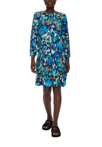 Buntbedrucktes Blumenkleid in Blau-, Grün- und Brauntönen; lange Ärmel, lockere Passform, geraffte Taille, kombiniert mit schwarzen Riemensandalen.