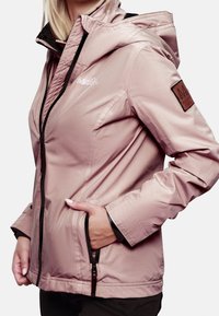 Femme portant une veste de plein air à capuche rose pâle avec des poches zippées, une main dans une poche, et des écussons de logo sur la poitrine et la manche.