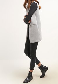 Szara kamizelka bez rękawów o gładkiej fakturze, pod spodem czarny długi rękaw, do tego czarne dopasowane dżinsy i botki za kostkę.