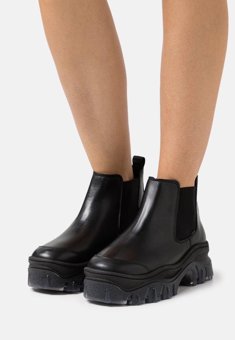 Jambes portant des bottines en cuir noir avec des semelles épaisses et robustes et des panneaux latéraux élastiques, sur un fond uni clair.
