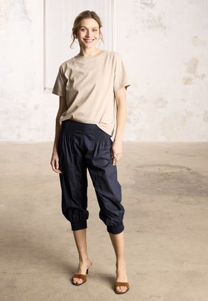 Smilende kvinde står indendørs iført en beige t-shirt, marineblå korte elastik-bukser og brune sandaler med hæl på en betongulv.