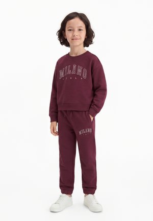 Kind mit schulterlangem Haar steht und trägt einen passenden bordeauxfarbenen Sweatshirt und Jogginghose mit dem Schriftzug „Milano Italy“ und weiße Sneakers.