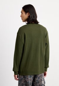 Olive zaļa ribota džemperis ar brīvu piegriezumu, nolaistām plecu daļām un garām piedurknēm, kopā ar kamuflāžas raksta kravas šortiem.