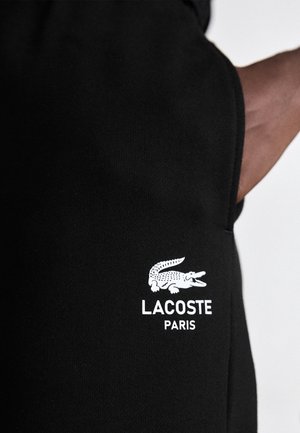 Czarna tkanina z białym logo Lacoste Paris przedstawiającym krokodyla, pokazana na górnej części ramienia lub okolicy ramienia osoby.