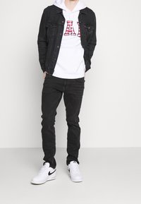 Veste en denim noire portée sur un sweat à capuche blanc avec des lettres rouges, associée à un pantalon noir et des baskets blanches avec un logo swoosh noir.