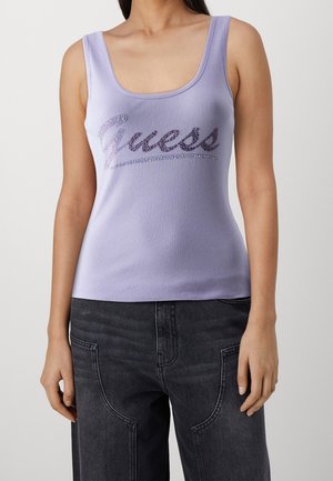 Nainen, joka pukeutunut laventelinsävyiseen, hihattomaan ribbattuun toppiin, jossa on strassikivillä koristeltu "Guess"-logo, yhdistettynä tummanharmaisiin korkeavyötäröisiin farkkuihin.