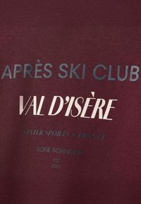 Sudadera burdeos con texto en blanco y verde azulado: "CLUB APRÈS SKI VAL D'ISÈRE DEPORTES INVERNALES • FRANCIA SOFIE SCHNOOR EST 2001." Textura de tela suave.