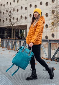 Modrý látkový batoh s šedými detaily, obdélníkový design, horní ucha a nastavitelné popruhy, drží osoba v jasně oranžové bundě a černých kalhotách.