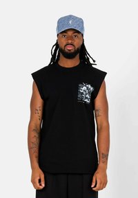 Maglietta nera senza maniche con un design grafico sul petto sinistro, realizzata in cotone. Indossata con un cappellino blu a motivo. Stile minimalista.