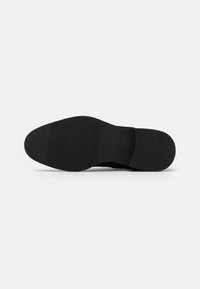 Semelle de chaussure noire avec une finition lisse, présentant un motif de grip texturé. Le design est plat avec un bout arrondi.