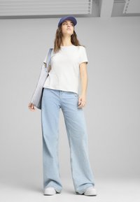 Camiseta de algodón gris claro, jeans de mezclilla de pierna ancha azul claro de corte holgado y una gorra lavanda. El modelo sostiene una bolsa de tela gris.