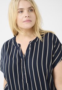 Blouse bleu marine à manches courtes avec des rayures verticales beiges, dotée d'une encolure à trou de bouton et d'un tissu lisse et léger.