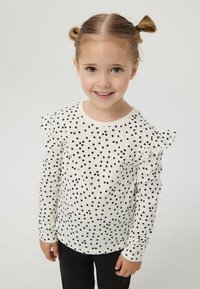 Biała bluzka z długim rękawem w czarne sercowate kropki, z falbaniastymi ramionami. W połączeniu z czarnymi spodniami, noszona przez dziecko.