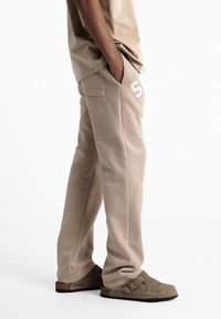 Pantalon de survêtement en polaire beige avec deux poches latérales, une coupe ample et un accent numérique blanc sur la cuisse droite. Texture douce, design simple.