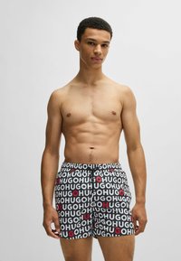 Black swim shorts with repeating "HUGO" text in white and red. Elastic waistband, modern cut. Model is shirtless with a neutral background.