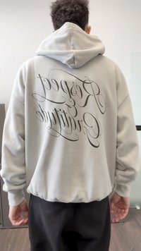 Pessoa vestindo um hoodie cinza claro com capuz, calças pretas, de pé dentro de casa com as costas voltadas para a câmera.