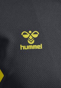 Hummel-Logo in Gelb auf strukturiertem schwarzen Stoff, mit einer Biene und dem Markennamen "hummel" in einer fetten Schriftart.